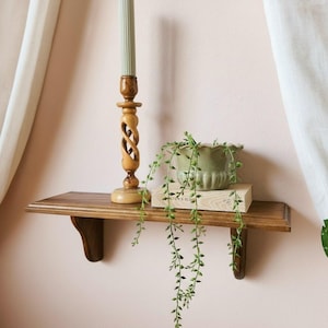 May include: A wooden wall shelf with two brackets holds a tall, light-colored fluted candle in a carved wooden holder, a light green scalloped ceramic planter, and a trailing string of pearls plant resting on a rectangular wooden block.