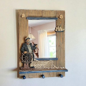 May include: A nautical-themed wall mirror with a weathered wood frame. The mirror features a sculpted captain figure, a ship's wheel, and a fishing net. Includes three blue hooks for hanging items. Decorative rope accents complete the design.