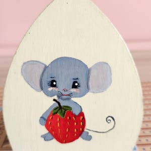 May include: A hand-painted, whimsical design featuring a light blue mouse holding a large red strawberry. The mouse has large ears and a curly tail. The background is a cream color.