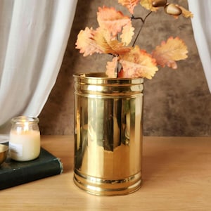 May include: A cylindrical polished brass vase with a reflective metallic surface. Inside the vase are several artificial autumn-colored oak leaves in shades of orange, yellow, and brown, featuring detailed veining and serrated edges on the foliage.