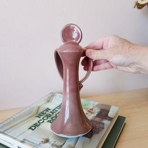 May include: A tall, mauve-colored ceramic vase with a unique design. The vase features a rounded top, a slender neck, and two curved handles. The vase is displayed on top of a stack of books.