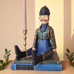 May include: A wooden figurine of a man in a plaid jacket, blue dress, and black boots, seated on books. The figurine has a mustache, monocle, and a black hat. A gold candlestick with a pink candle is to the left.