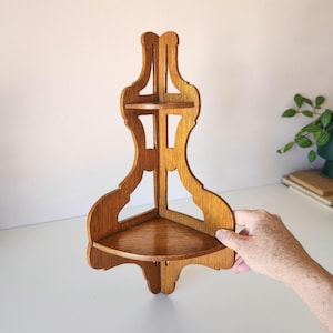 May include: A light brown wooden corner shelf featuring two tiers with a cutout design. The structure is composed of vertical panels that meet at a right angle, supporting two triangular shelves with a smooth, polished finish and visible wood grain.