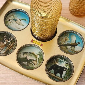 May include: A vintage gold-toned metal serving tray with six circular wildlife illustrations. The illustrations include a duck, deer, fish, pheasant, and bear. A textured amber glass sits in the center. The tray has a label that reads "Souvenir of Black Van Tassell".