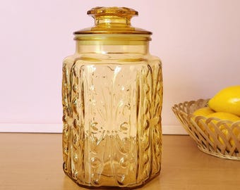 May include: A vintage amber glass jar with a lid. The jar has a rectangular shape with embossed floral patterns. The lid is also amber-colored and has a decorative knob. The jar is likely used for storing food items.