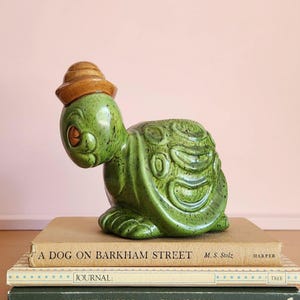 May include: A green ceramic turtle figurine wearing a brown hat. The turtle has a speckled finish and detailed shell design. The figurine is sitting on top of a stack of books with the title "A Dog on Barkham Street".