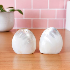 May include: A set of two white mother of pearl salt and pepper shakers. The shakers are egg-shaped and have a pearlescent finish.