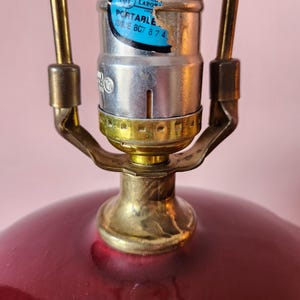 May include: Close-up of a vintage lamp with a deep red, rounded base and brass-colored metal accents. The lamp's socket is silver with a label that reads "PORTABLE ISSUE BC7 874". The lamp's design suggests a classic or retro style.