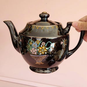 May include: A black ceramic teapot with a lid and handle. The teapot is decorated with a floral design in shades of yellow, blue, pink, and green. Gold trim accents the lid and rim. The teapot is held by a hand.