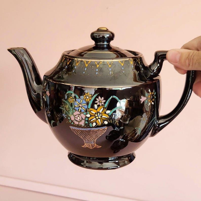 May include: A black ceramic teapot with a lid and handle. The teapot is decorated with a floral design in shades of yellow, blue, pink, and green. Gold trim accents the lid and rim. The teapot is held by a hand.