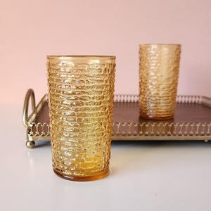 May include: Two amber glass tumblers with a textured surface sit on a gold metal tray with a brown wood base.