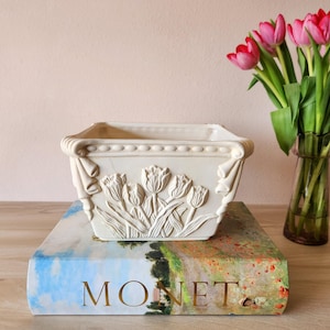 May include: Cream-colored rectangular planter with embossed tulip design. The planter sits atop a book with the word "MONET" visible. A vase of pink tulips is in the background.