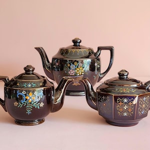 May include: Three antique teapots in a row. The teapots are dark brown with floral and geometric designs in various colors. The teapots have gold accents and ornate handles and spouts. The background is a solid pink.