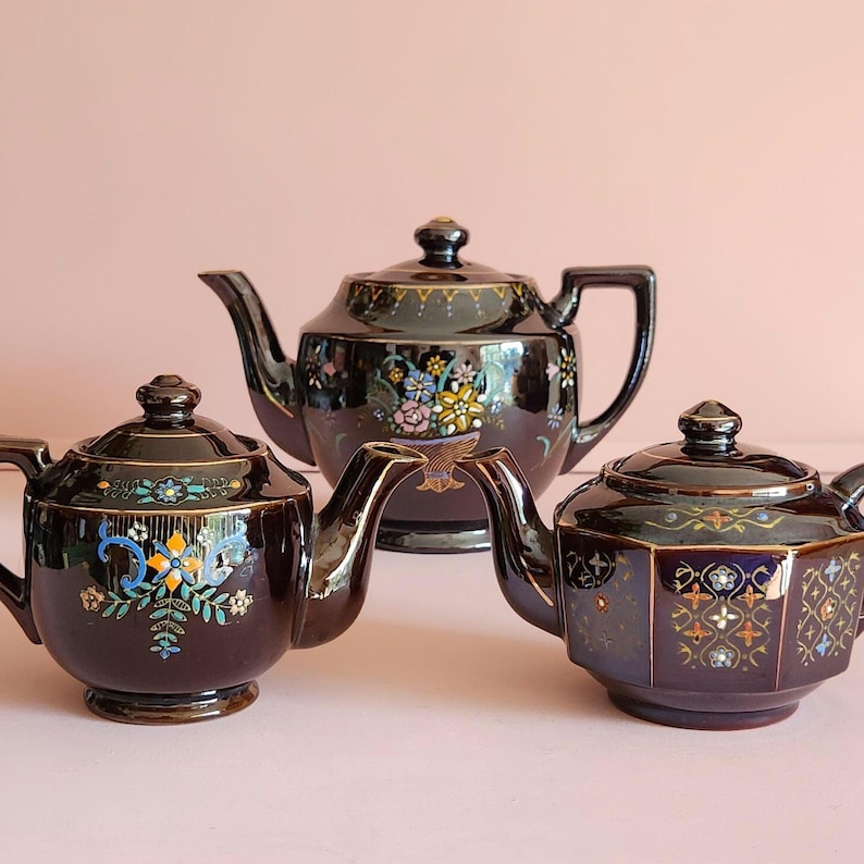 May include: Three antique teapots in a row. The teapots are dark brown with floral and geometric designs in various colors. The teapots have gold accents and ornate handles and spouts. The background is a solid pink.
