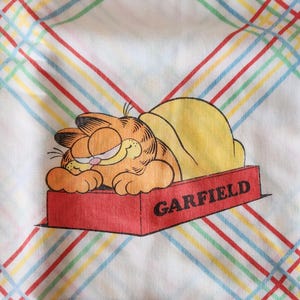 May include: A vintage sheet featuring Garfield the cat, lying in a red box with the name "GARFIELD" printed on it. The sheet has a white background with a plaid pattern of red, green, yellow, and blue lines.