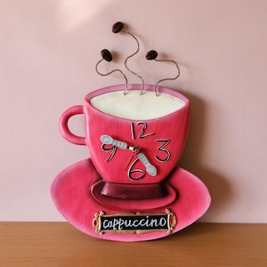 May include: Pink ceramic coffee cup clock with a white face and black numbers. The clock has a red second hand and a silver hour and minute hand. The cup has a saucer and a sign that says "cappuccino".