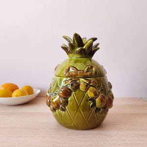 May include: A green ceramic pineapple-shaped jar with a lid, adorned with brown fruit and bow details. The jar has a diamond pattern and a textured surface. The jar is on a wooden surface.