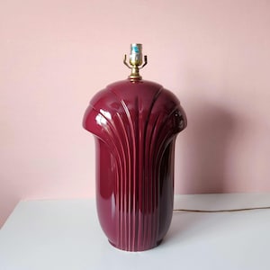 May include: A burgundy-colored ceramic table lamp with a fluted design. The lamp has a rounded top and a cylindrical base. The lamp has a brass-colored light fixture and a brown electrical cord. The lamp is on a white surface against a pink background.