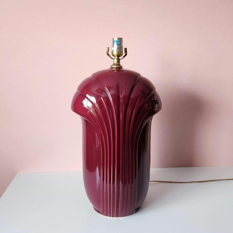 May include: A burgundy-colored ceramic table lamp with a fluted design. The lamp has a rounded top and a cylindrical base. The lamp has a brass-colored light fixture and a brown electrical cord. The lamp is on a white surface against a pink background.
