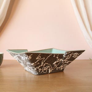 May include: A square ceramic bowl with a dark gray, speckled exterior featuring white, abstract, drip-style glaze patterns. The interior of the bowl is a smooth, light mint green color, and the rim has a distinct, angular, flared shape.