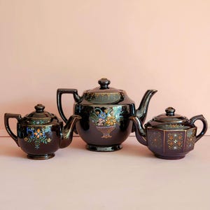May include: Three antique black teapots with gold trim and floral designs. The teapots vary in size, with the largest featuring a basket of flowers. The teapots are arranged on a light pink surface.