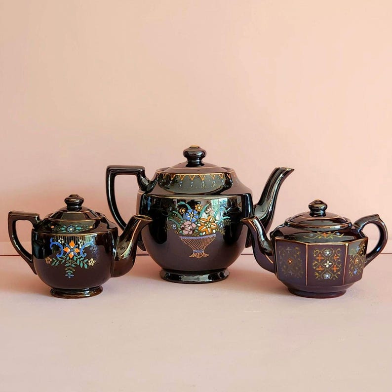 May include: Three antique black teapots with gold trim and floral designs. The teapots vary in size, with the largest featuring a basket of flowers. The teapots are arranged on a light pink surface.