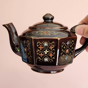 May include: A dark brown ceramic teapot with a lid and handle. The teapot has a geometric design with floral patterns in red, blue, and gold. The teapot is held by a hand.