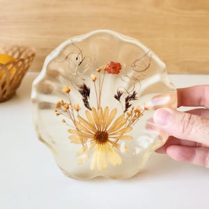 May include: A circular, scalloped-edge resin coaster featuring embedded dried flowers, including a large yellow daisy, small yellow sprigs, dark brown grasses, and a single red bud, all preserved within a clear, glossy, transparent material.