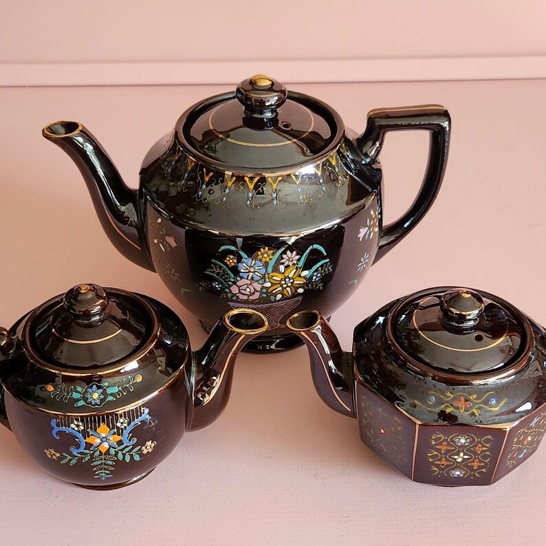 May include: Three black ceramic teapots with gold trim and floral designs. The largest teapot has a rounded body and a curved handle. The smaller teapots have a more compact design. The teapots are arranged on a pink surface.