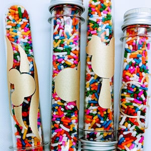 May include: Four clear glass tubes filled with colorful sprinkles. Each tube has a gold sticker with a different design. The tubes are on a white surface.