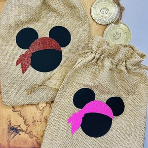 May include: Two burlap gift bags with a Mickey Mouse design. The bags are decorated with black ears, a black face, and a red or pink glitter bandana. The bags are sitting on a brown surface.
