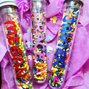 May include: Three clear plastic test tubes filled with colorful sprinkles. Each tube has a name on it: "Megan", "Grace", and "Javier".