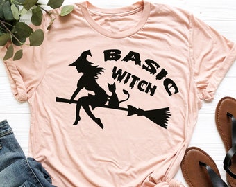 May include: A pink t-shirt with a black silhouette of a witch riding a broomstick with a black cat. The text "BASIC WITCH" is written in black above the witch. The t-shirt is on a white surface with a pair of brown sandals and blue jeans.
