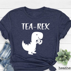 May include: A navy blue t-shirt with a white graphic of a dinosaur holding a teacup. The text "TEA-REX" is printed above the dinosaur.