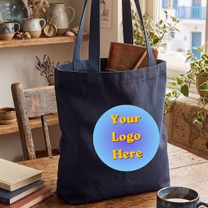 May include: A dark navy blue canvas tote bag featuring a circular light blue gradient patch on the front with the yellow text "Your Logo Here" printed in the center. The bag has two long fabric handles and is standing upright on a wooden surface.