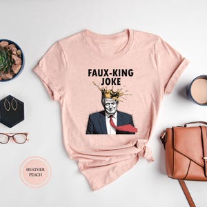 May include: A light pink t-shirt with the text "FAUX-KING JOKE" above an illustration of a man wearing a crown. The shirt is surrounded by accessories, including a brown purse, glasses, and a succulent.