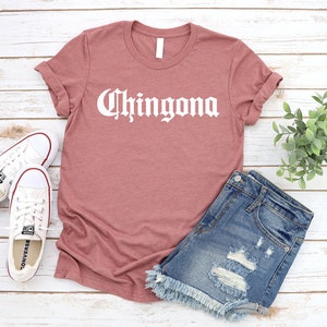 May include: A mauve t-shirt with the word "Chingona" printed in white on the front. The shirt is styled with denim shorts and white sneakers.