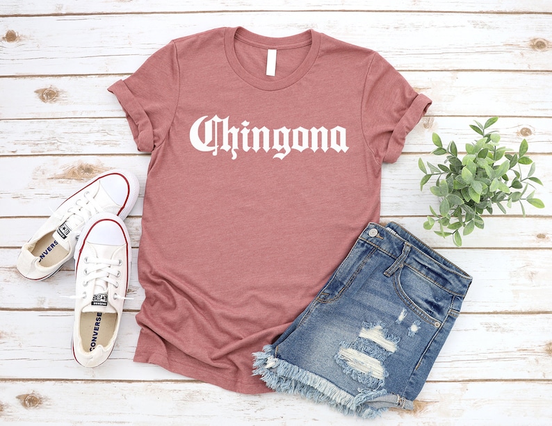 May include: A mauve t-shirt with the word "Chingona" printed in white on the front. The shirt is styled with denim shorts and white sneakers.