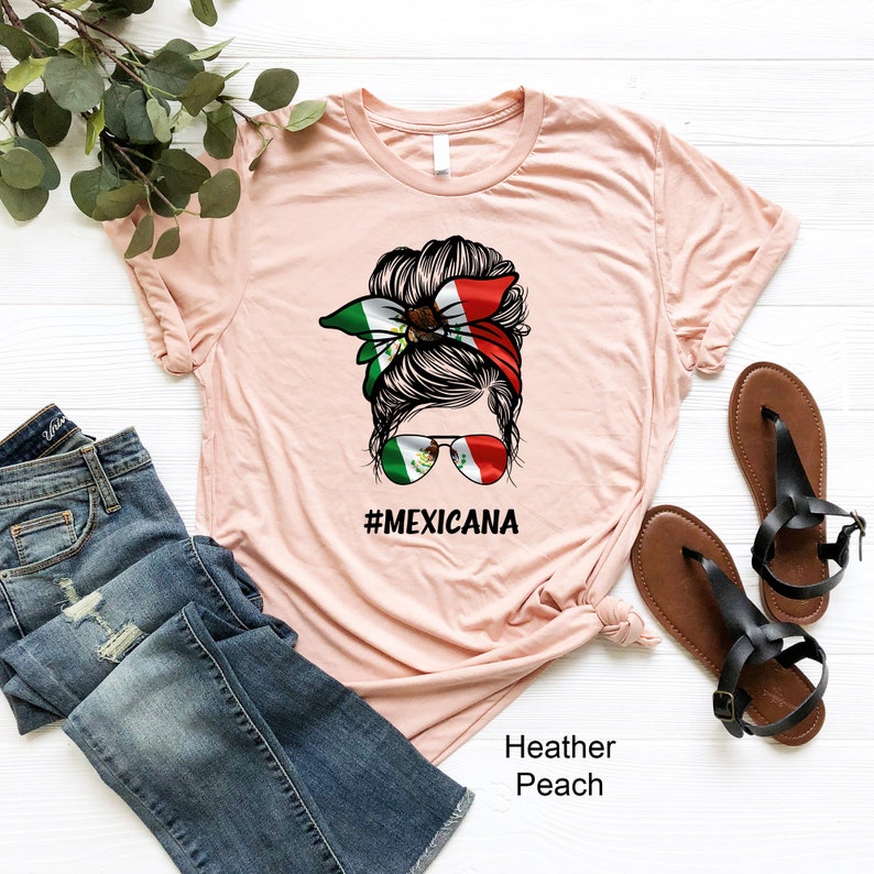 May include: A peach colored t-shirt with a graphic of a woman with a messy bun wearing a bandana with the Mexican flag colors and sunglasses with the Mexican flag on them. The text "#MEXICANA" is printed below the graphic. The t-shirt is on a white surface with a pair of brown sandals and blue jeans.