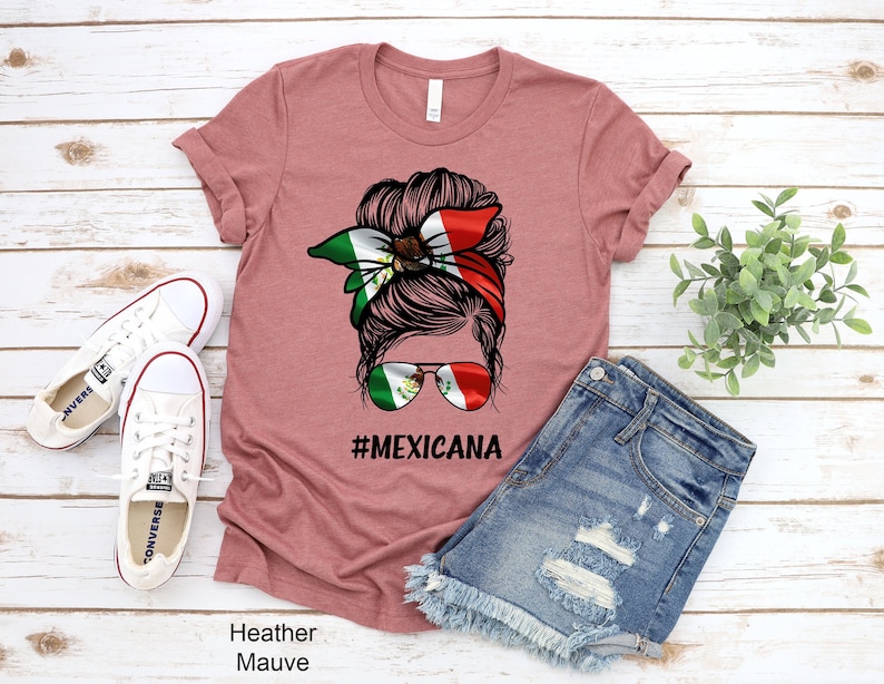 May include: A heather mauve t-shirt with a graphic of a woman with a messy bun wearing sunglasses and a headband with the Mexican flag. The text "#MEXICANA" is printed below the graphic.