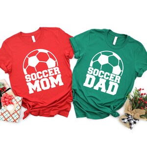 May include: Two t-shirts, one red and one green, with a white soccer ball graphic and the text "Soccer Mom" and "Soccer Dad" in white.