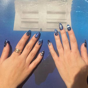 Op de afbeelding: Handen met lange, amandelvormige nagels. De nagels zijn geschilderd in verschillende blauwtinten, met ontwerpen zoals sterren, een dambordpatroon en gedetailleerde illustraties. Een gouden ring siert een hand.
