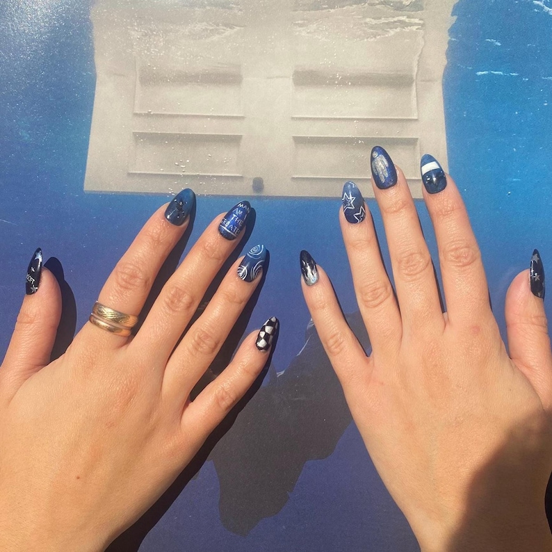 Op de afbeelding: Handen met lange, amandelvormige nagels. De nagels zijn geschilderd in verschillende blauwtinten, met ontwerpen zoals sterren, een dambordpatroon en gedetailleerde illustraties. Een gouden ring siert een hand.