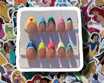 Op de afbeelding: Een set kunstnagels met ontwerpen geïnspireerd op Disney-prinsessen. Elke nagel heeft een nude basis met kleurrijke accenten, waaronder geel, blauw, roze en groen. Sommige nagels hebben kleine versieringen zoals een kroon of een hart.