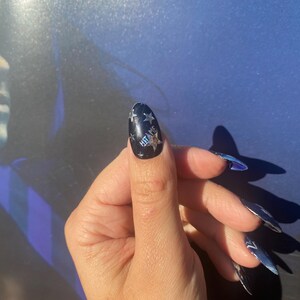 Op de afbeelding: Een hand met lange, puntige, blauwe en zilveren nagels. De duimnagel is zwart met zilveren sterontwerpen en de woorden "HIT ME". De nagels zijn een uniek en stijlvol accessoire.