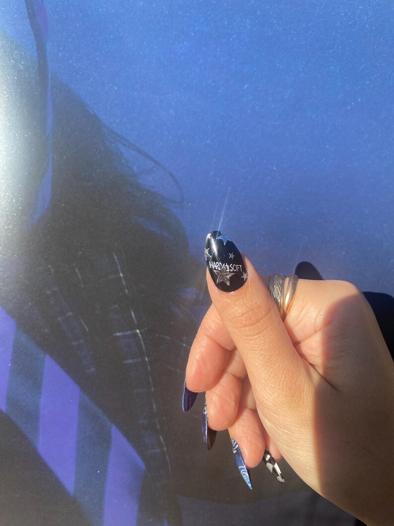 Op de afbeelding: Een hand met lange, donkerblauwe vingernagels. E&eacute;n nagel heeft de tekst "HARD & SOFT" met sterontwerpen op een zwarte achtergrond. Andere nagels hebben een dambordpatroon en een blauw-wit ontwerp.