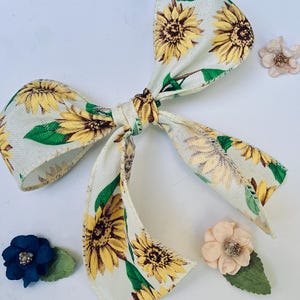 May include: A cream-colored bow with a sunflower pattern, featuring yellow sunflowers and green leaves. The bow is accompanied by three decorative flowers in shades of blue and peach.