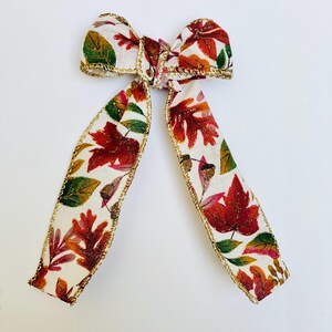 May include: A decorative bow featuring a fall leaf pattern in red, green, and gold on a cream background. The edges are trimmed with gold metallic thread. This bow is suitable for seasonal decor.