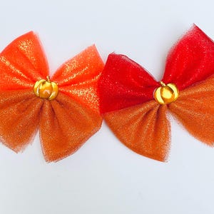 May include: Two decorative hair bows in shades of orange and red, each featuring a gold pumpkin charm in the center. The bows are made of a shimmering, sheer material, perfect for fall-themed accessories.