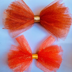 May include: Two orange tulle hair bows with gold and yellow ribbon centers. The bows are made of a shimmering, sheer fabric, and are arranged on a light blue surface. The bows are a vibrant, eye-catching accessory.